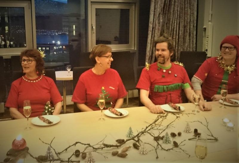 Fire personer kledt i rødt ved et bord dekket til julelunsj. Smil og god steming.